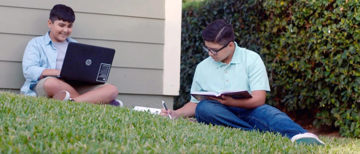 Two boys sit on grass near a house, one with a laptop and smiling, the other writing in a notebook. The scene is calm and focused; sunny day.